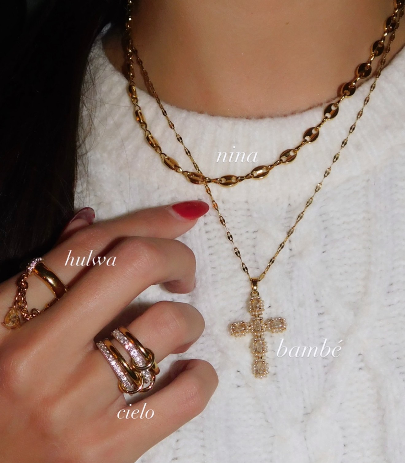 Close-up of a person wearing layered gold necklaces and rings, with a white sweater and red nail polish.