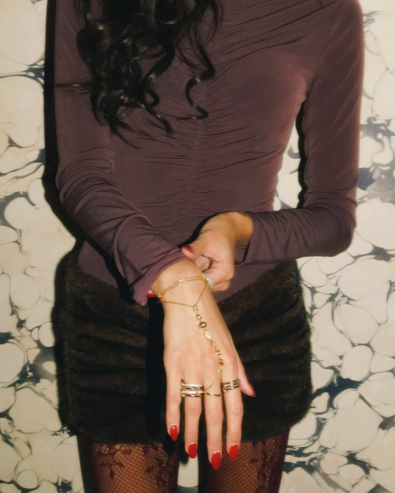 Person wearing a long-sleeve top, black skirt, and patterned tights, holding a gold bracelet and rings, with red nail polish.