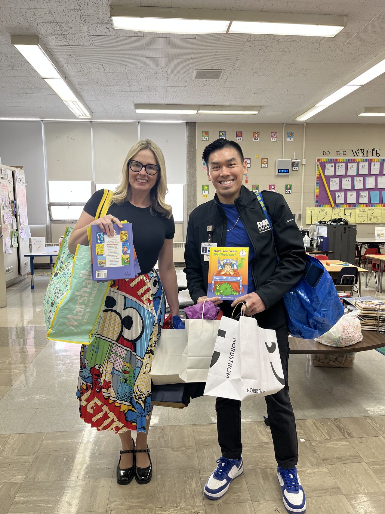 Two people standing in a classroom holding colorful books and bags, smiling at the camera.