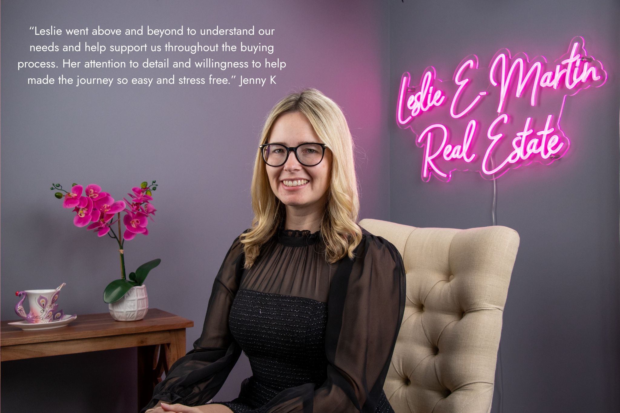 Woman with glasses and blonde hair sitting on a beige tufted chair, smiling, in front of a wall with pink neon sign and a quote.