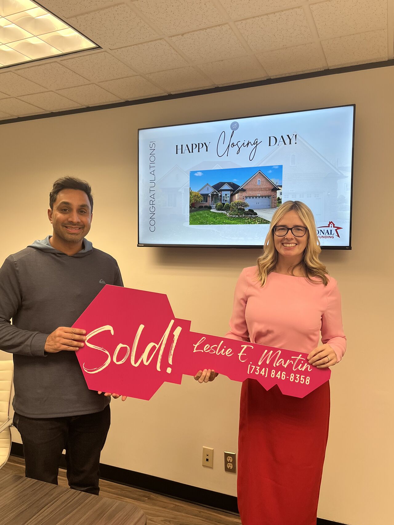 Man and woman holding a sold sign in a room with a large screen displaying a house and birthday message.
