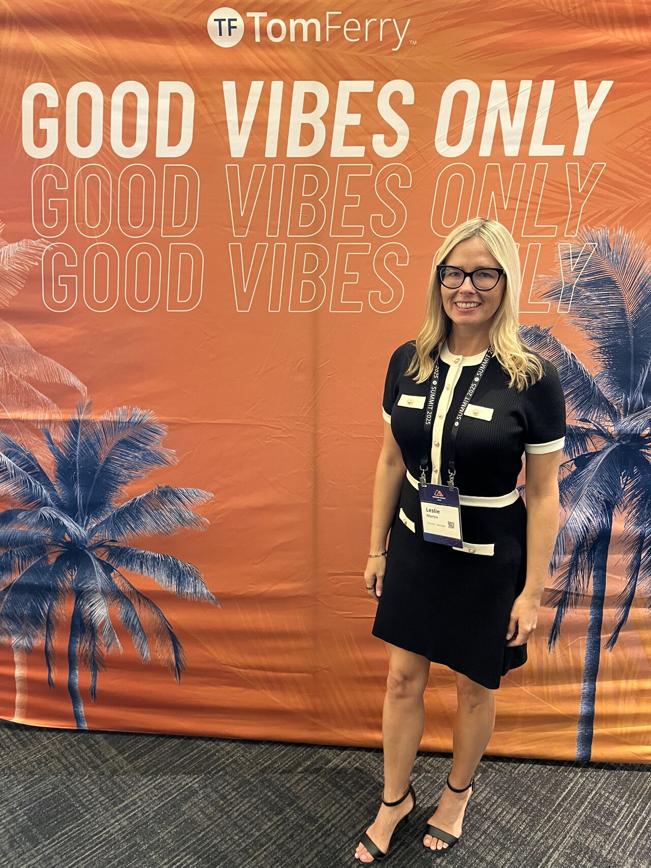Woman with glasses and blonde hair standing in front of a tropical-themed backdrop with palm trees and text.