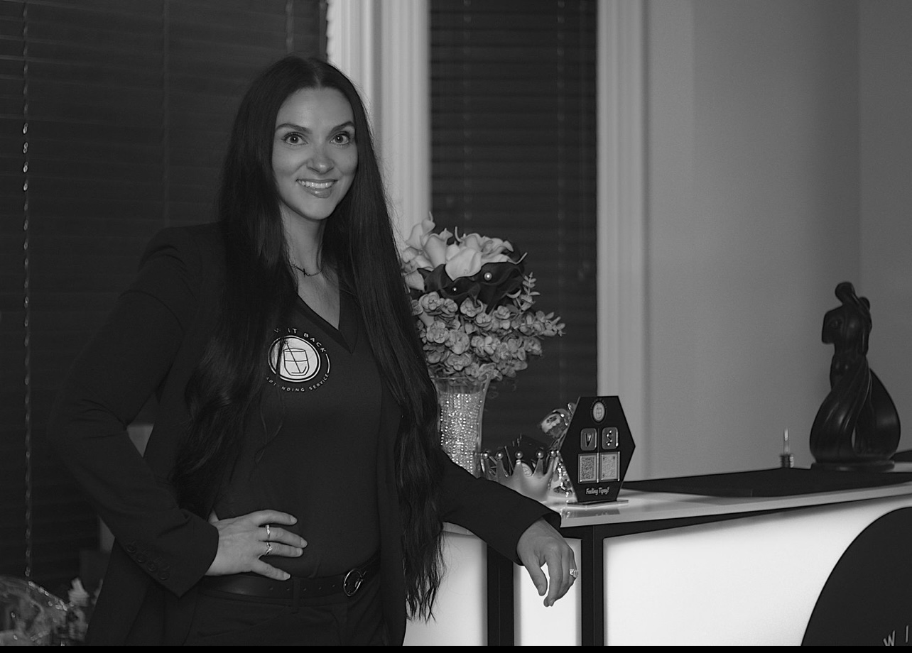 Woman with long dark hair standing indoors near a counter with a flower arrangement and decorative items.