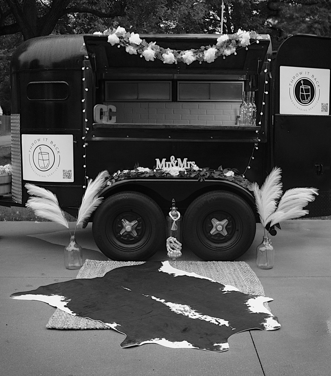 Decorated mobile food cart with flowers, a rug in front, and bottles on the ground, outdoors.