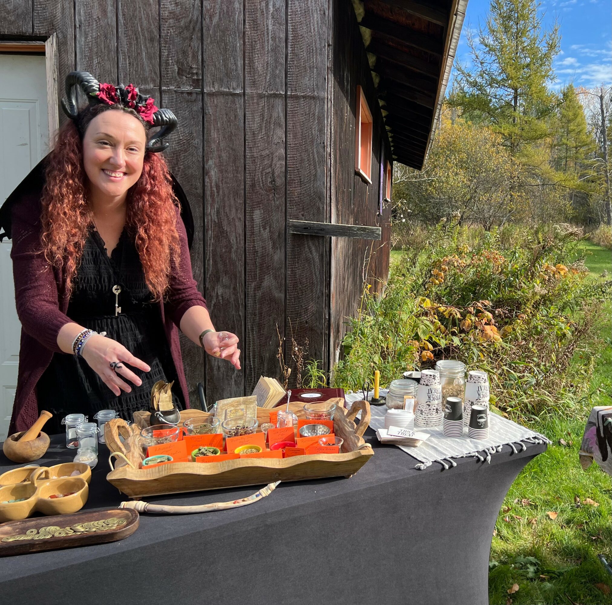 Woman with floral headband smiling at outdoor table with food and drinks, wooden building and trees in background.