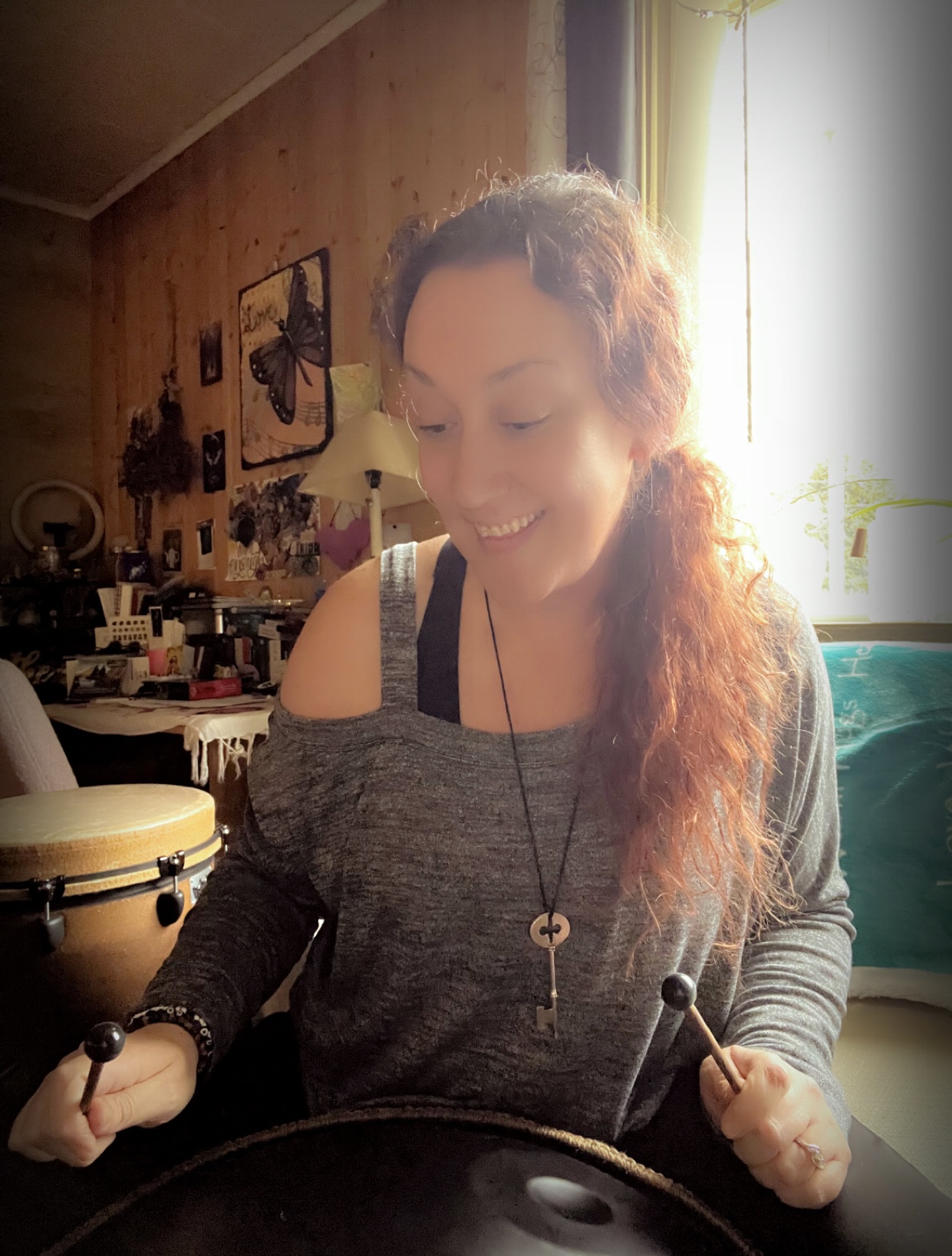 Woman with long, wavy hair smiling, sitting indoors near a window with sunlight, holding drumsticks.