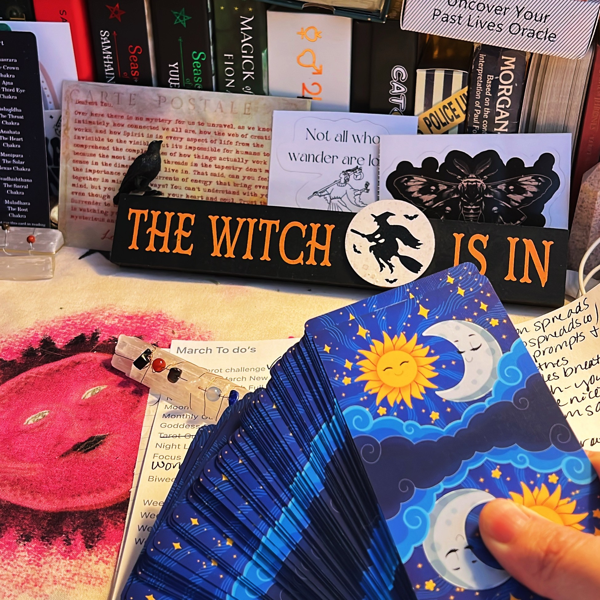 Hand holding a fan of colorful tarot cards with sun and moon illustrations, surrounded by books and a pink face mask.