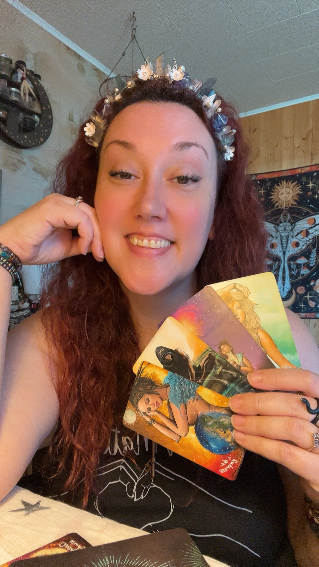 Woman with long curly hair wearing a floral headband, smiling, holding colorful tarot cards, sitting at a table.