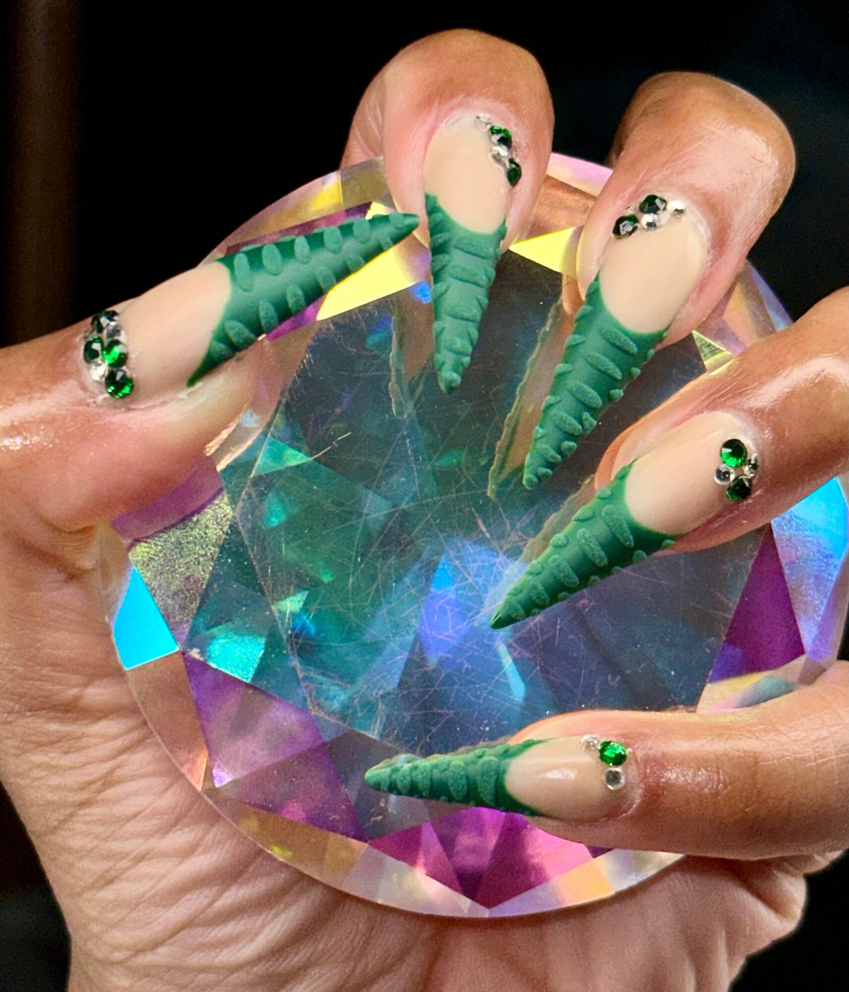 Hand with long, decorated nails holding a large, faceted, colorful crystal.