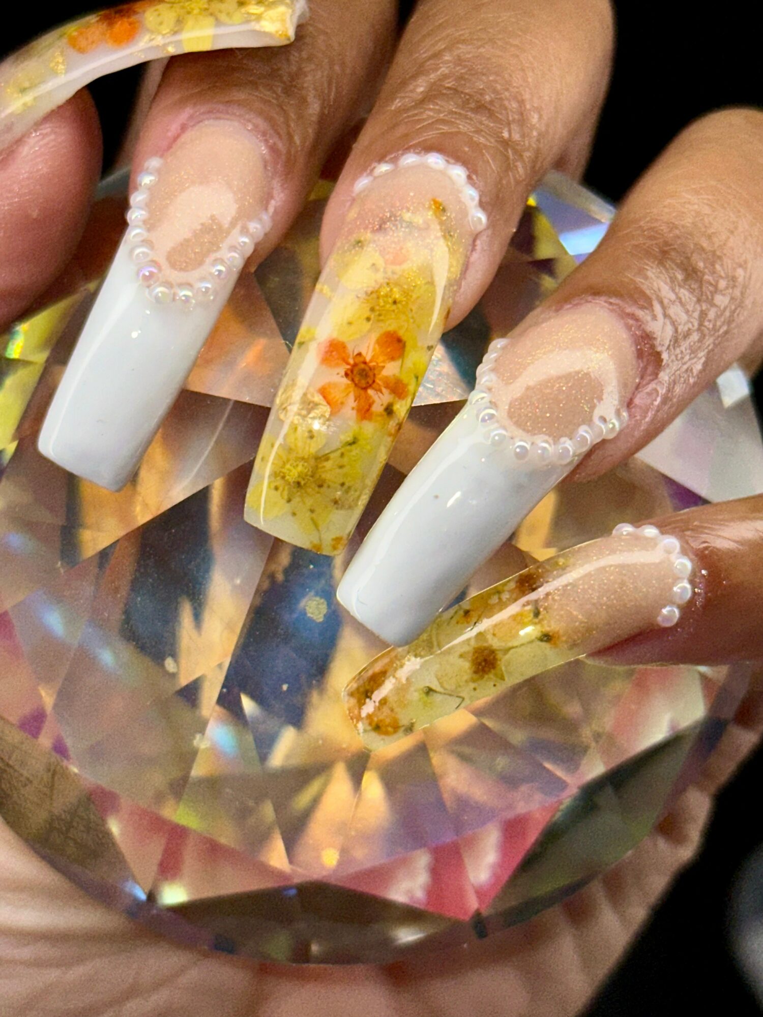 Close-up of five manicured nails with white and yellow floral designs, some with clear tips, on a reflective surface.