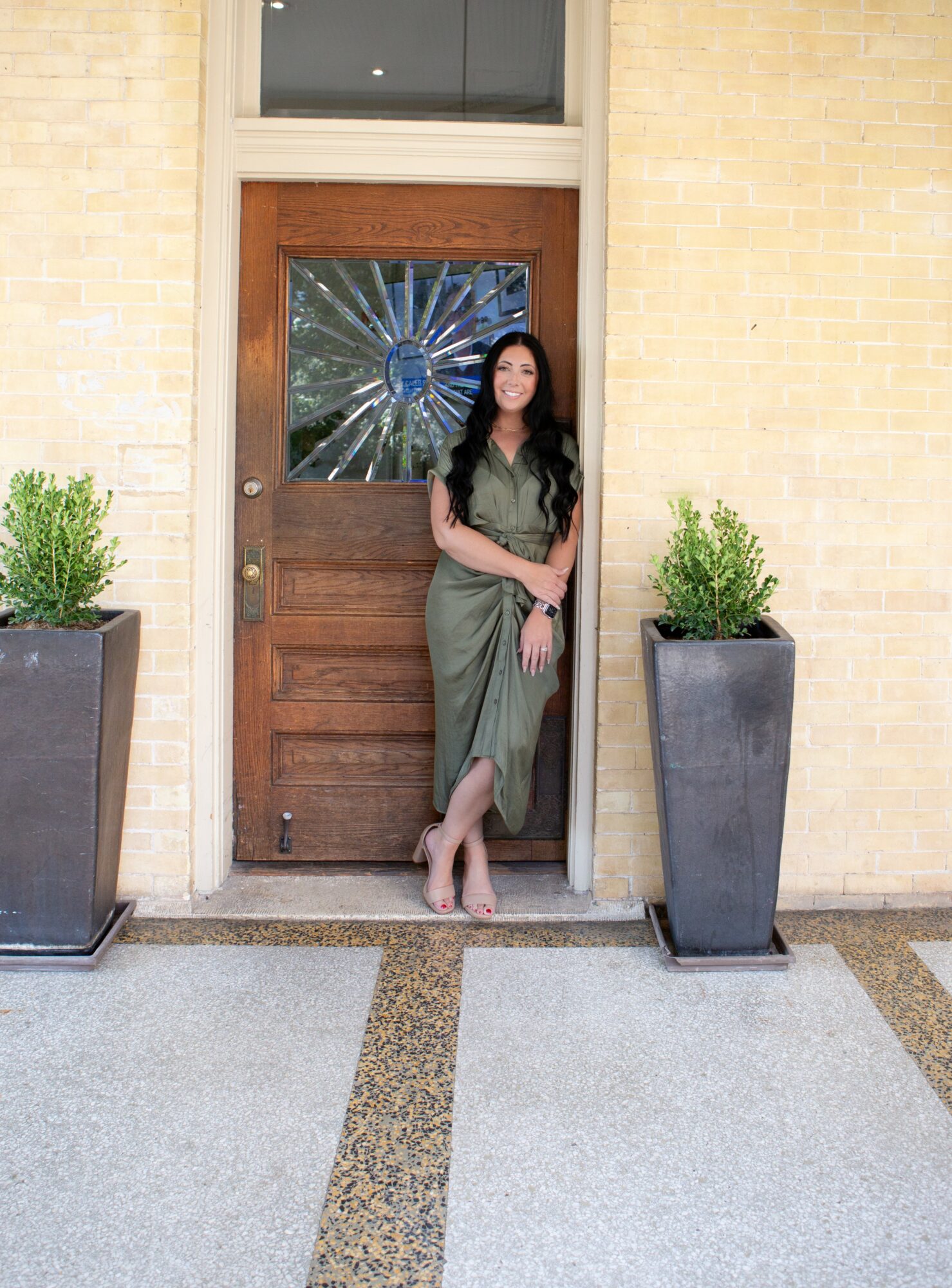 Woman in green dress standing in front of a wooden door with a shattered glass window, flanked by two large potted plants.