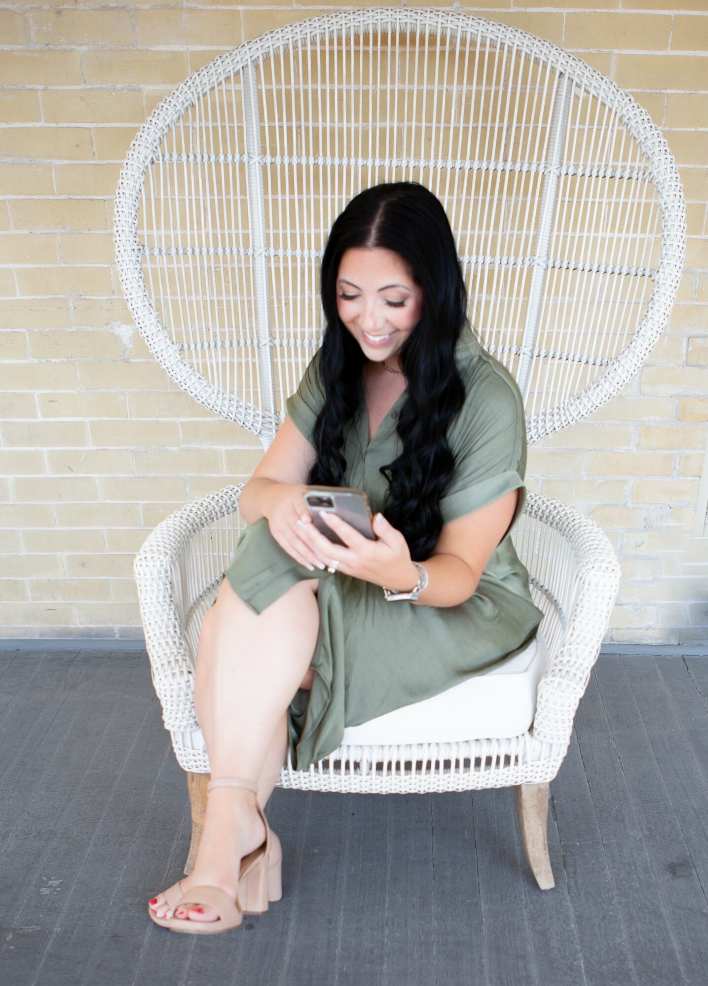 Woman with long dark hair sitting on a white wicker chair, looking at her phone, against a brick wall background.
