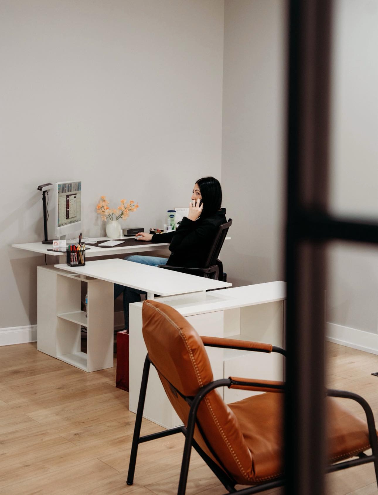 Person sitting at a desk in a modern office, viewed through a partially open door, with a brown chair in foreground.
