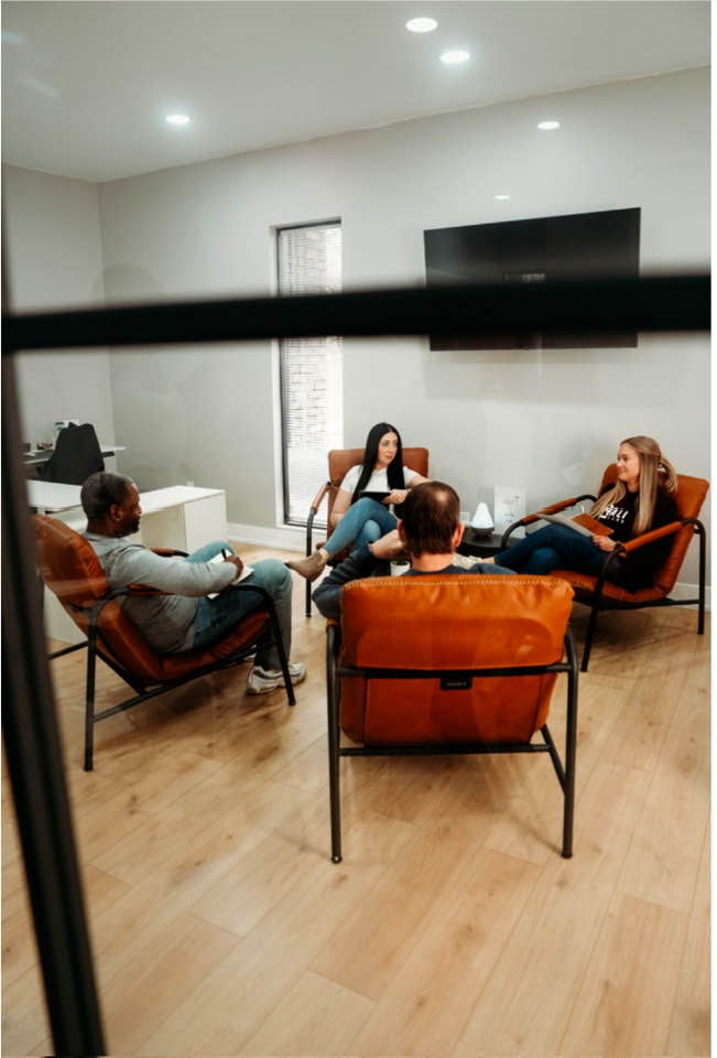 Four people sitting in a circle on chairs in a room with a large TV on the wall.