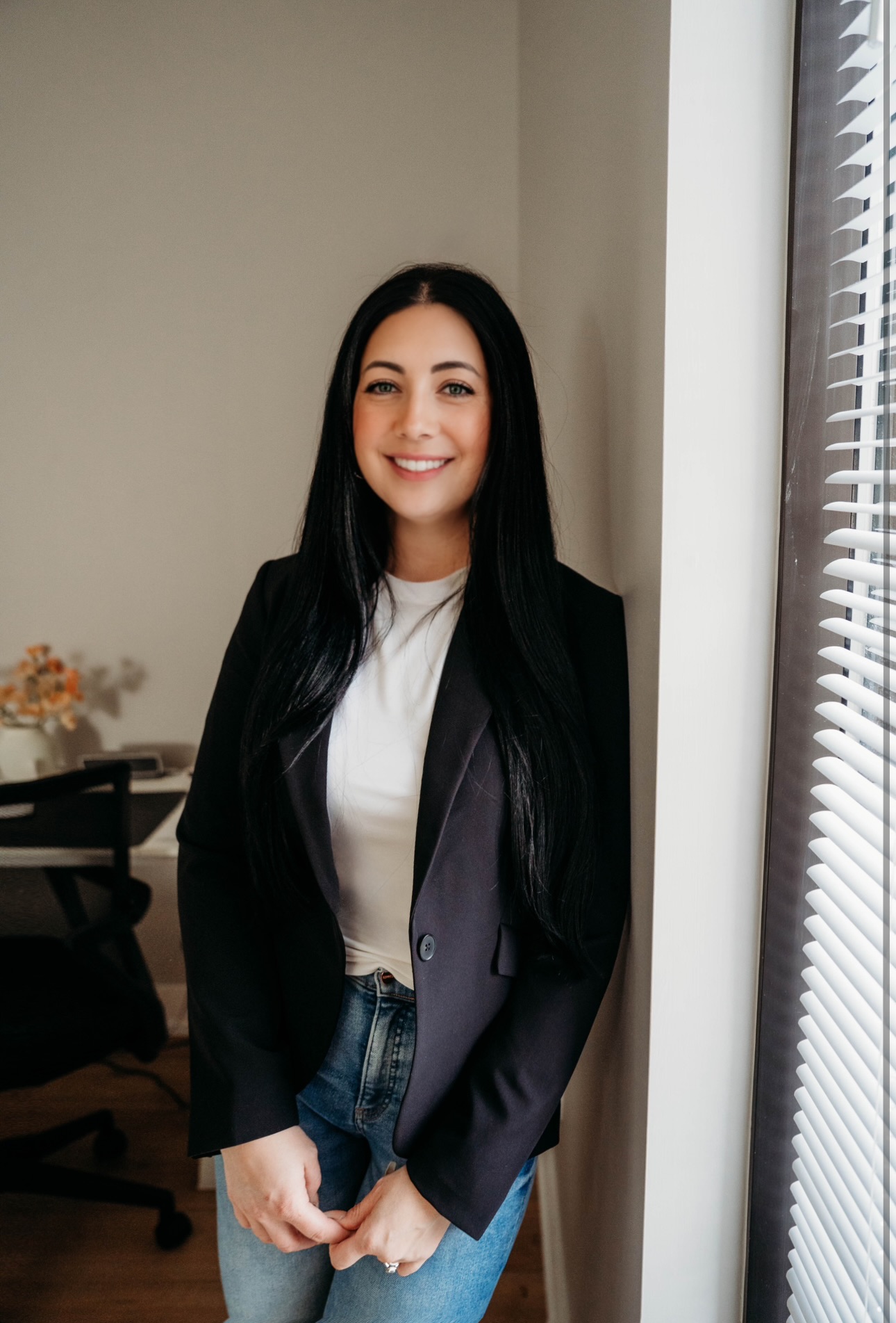 Smiling woman with long dark hair standing indoors near window with blinds, wearing a black blazer and white top.