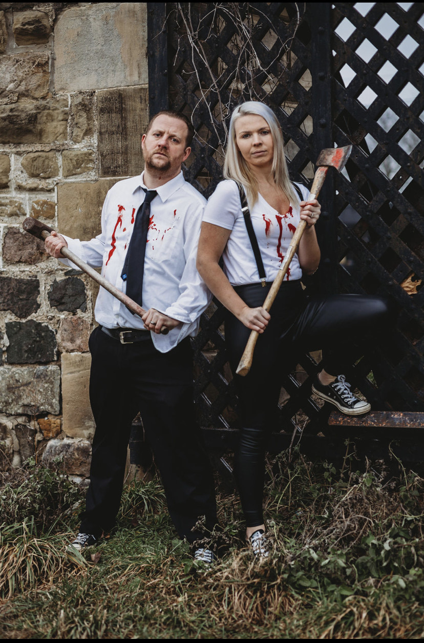 Man and woman stand outdoors near a stone wall and wooden structure, holding axes, dressed in casual clothes with bloodstains.