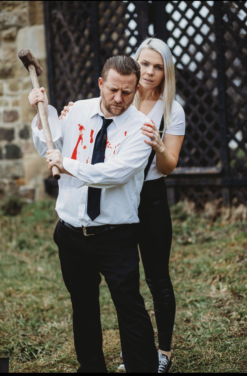 Man with bloodstained shirt holding axe, woman standing behind him outdoors, both with serious expressions.