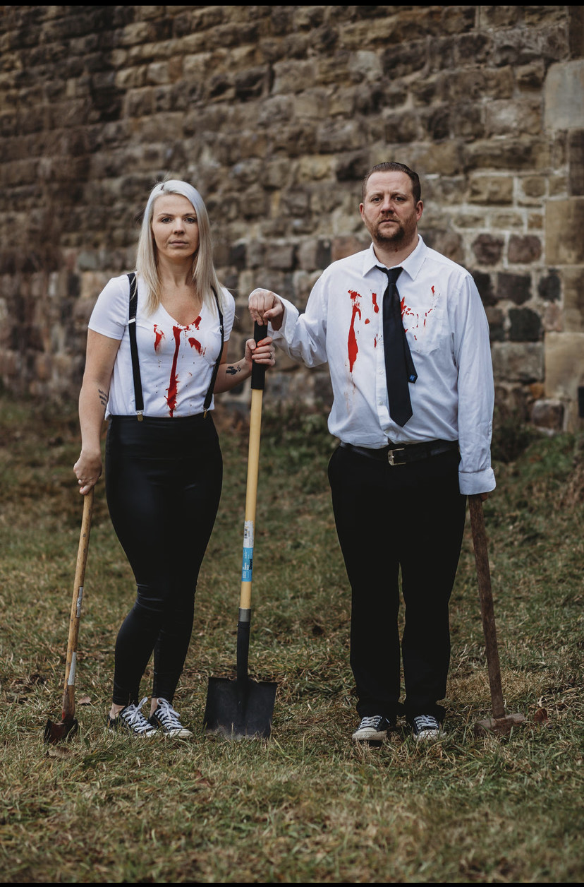 Two people stand outdoors against a stone wall, holding shovels with red paint on their clothes.
