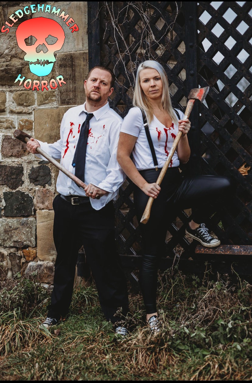 Two people in horror-themed costumes stand outdoors near a stone wall and wooden fence, holding axes, with a horror logo in the top left.