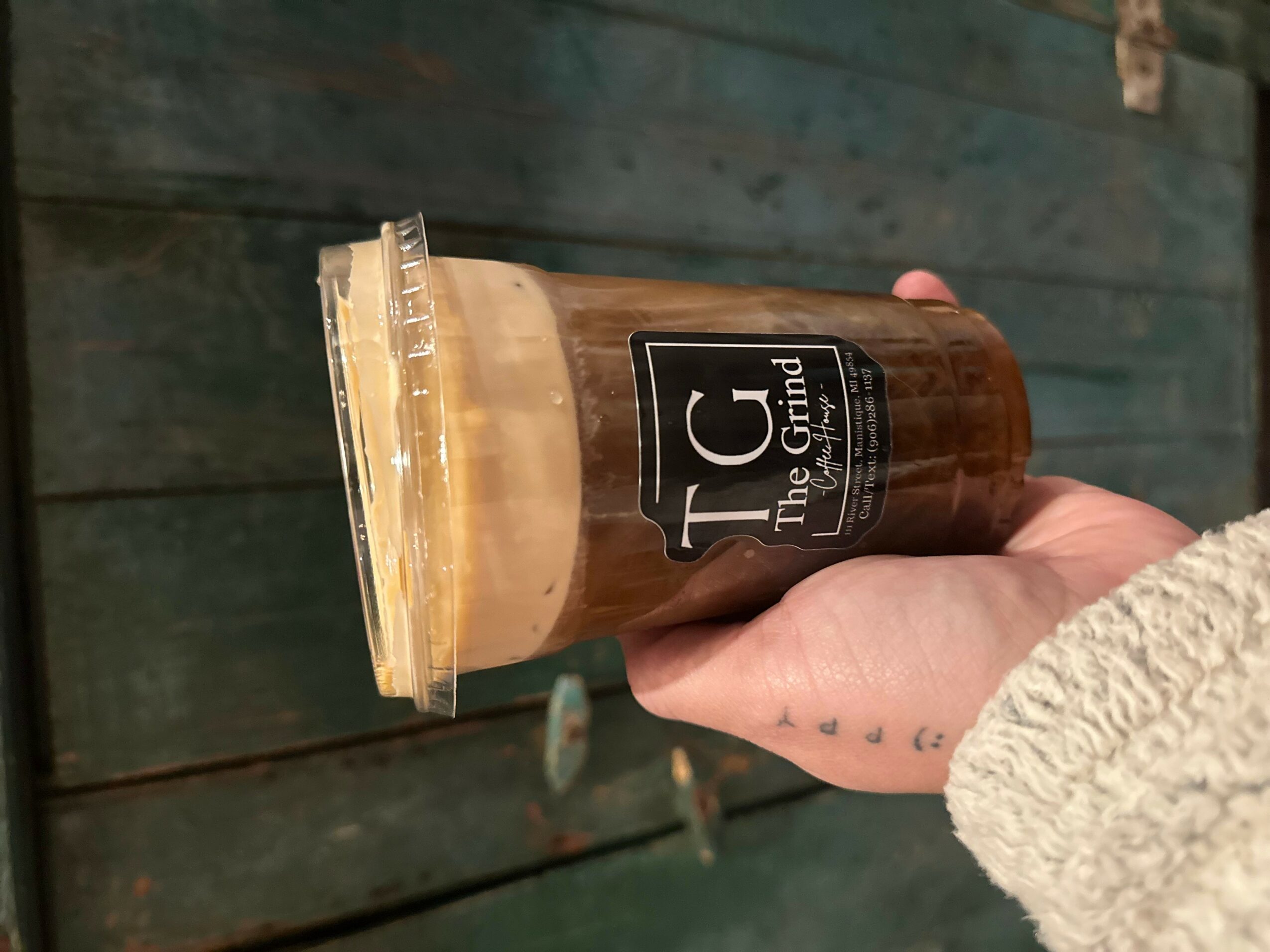 Hand holding iced coffee in a plastic cup with a black label, against a wooden background.