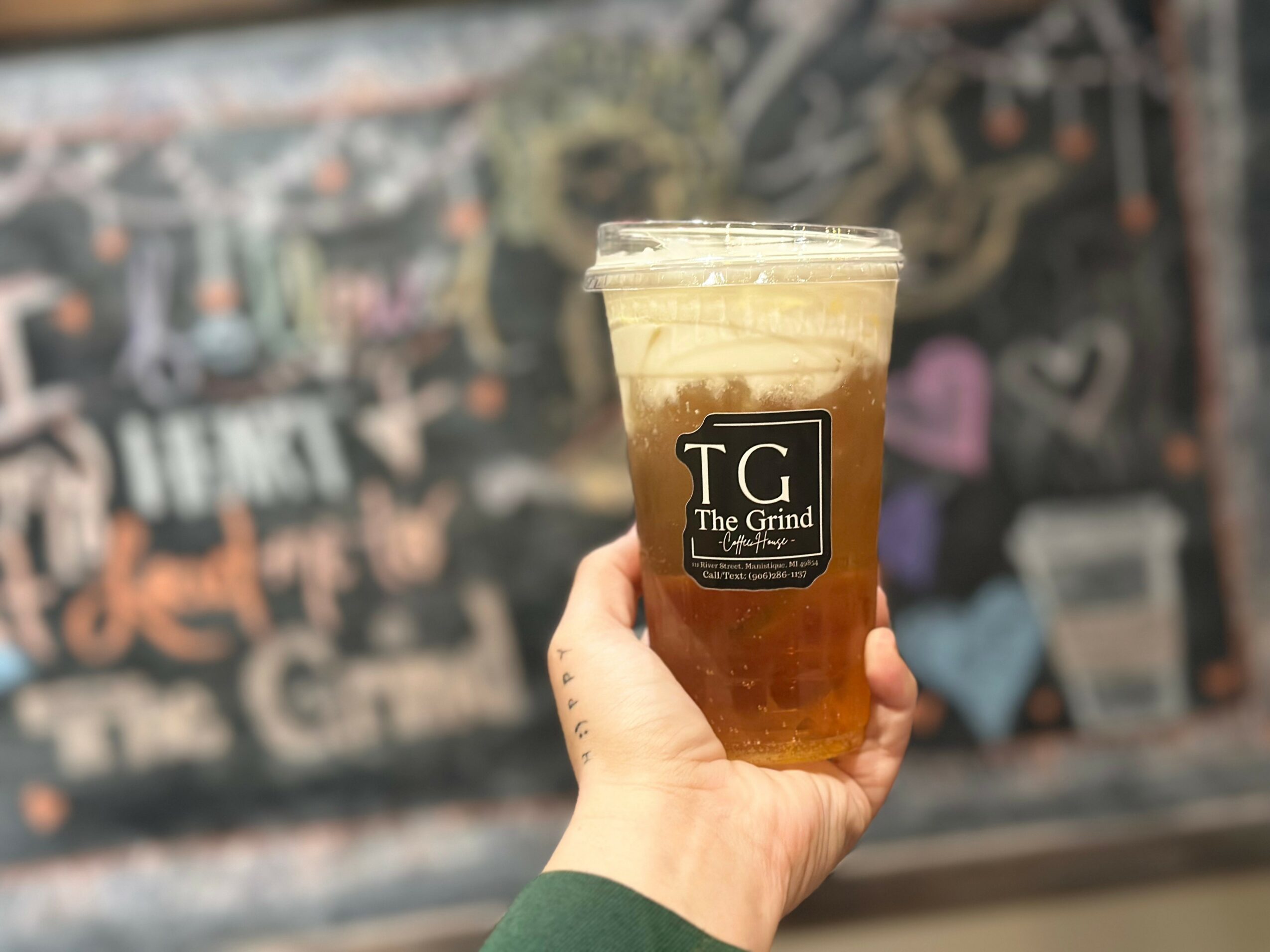 Hand holding a plastic cup with a dark beverage and foam on top, in front of a chalkboard wall with colorful drawings.