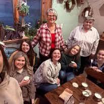 Group of seven people gathered around a table indoors, smiling, with plants and decorations in the background.