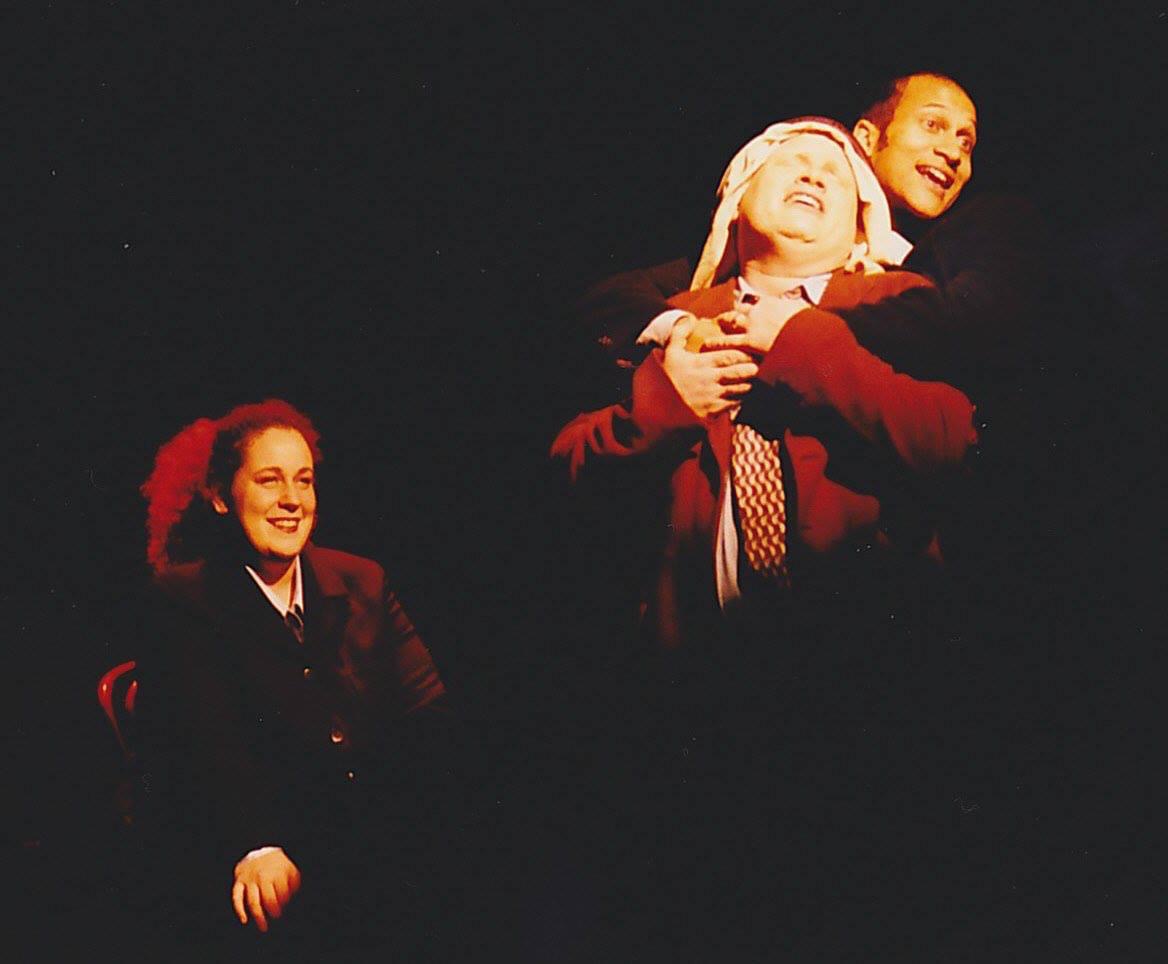 Three people on stage, two are laughing, one is smiling, dark background, theatrical setting.