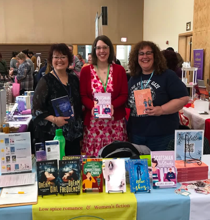 Three women stand behind a table with books at a book fair or event, smiling and holding books.