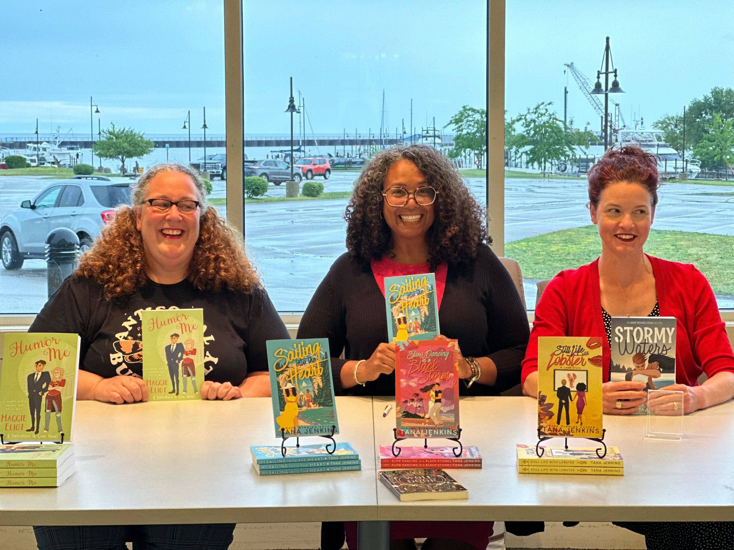 Three women sitting at a table with books displayed in front of them, large window with a parking lot and boats outside.