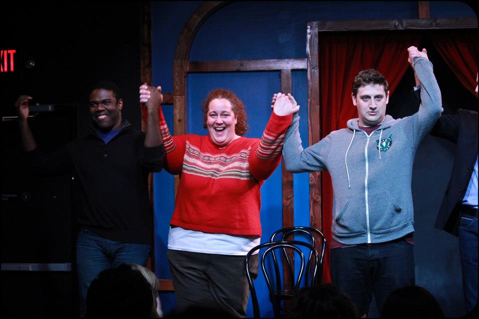 Three people holding hands and raising arms, smiling on a stage with a red curtain background.