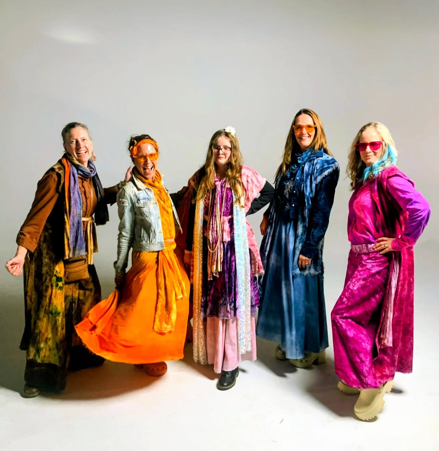 Five women stand together, wearing colorful, flowing outfits and scarves, smiling against a plain background.