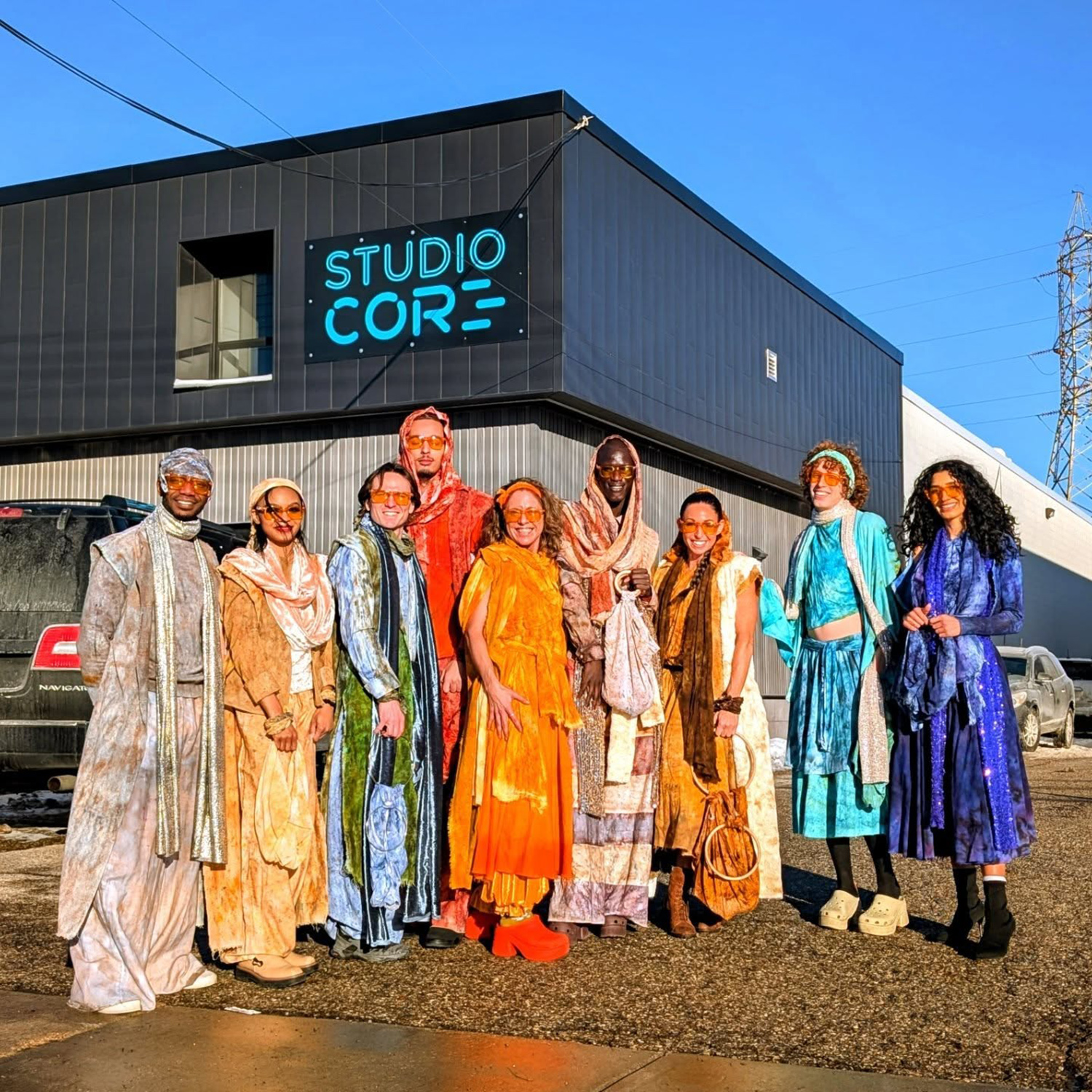 Group of ten people standing outside a building with a sign that reads 'STUDIO COR'. They are dressed in colorful clothing and wearing masks.
