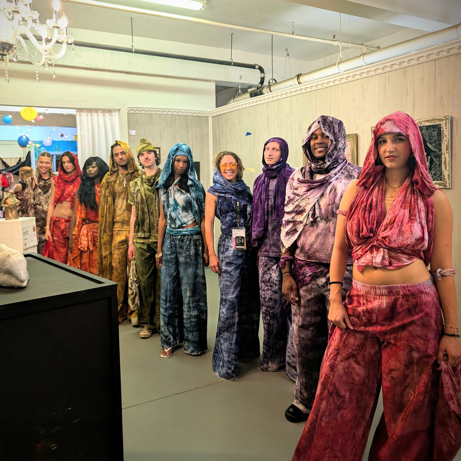 Line of people in colorful, textured clothing standing indoors with paintings on the wall.