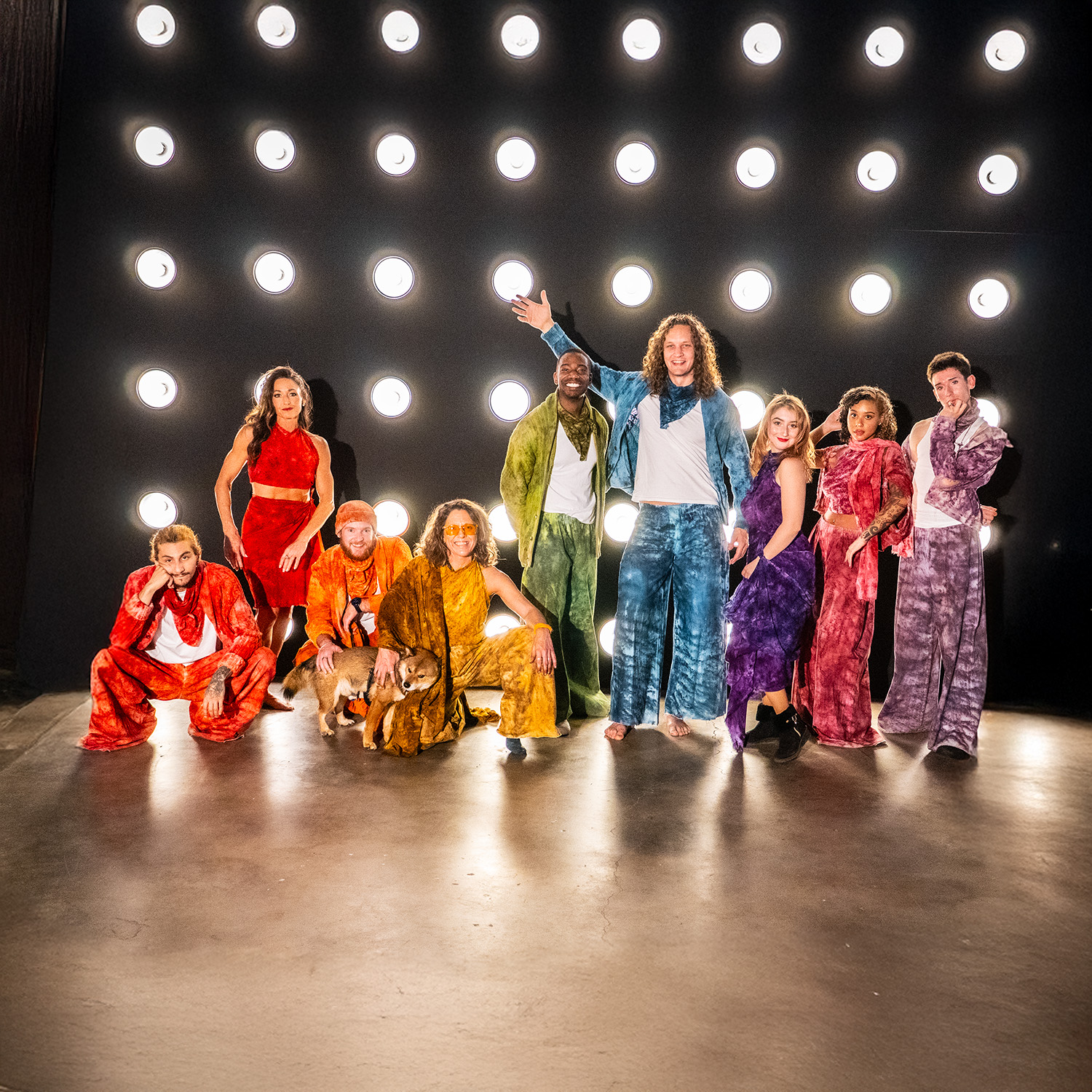 Group of nine people on stage in colorful outfits, some standing and some sitting, with bright lights behind them.