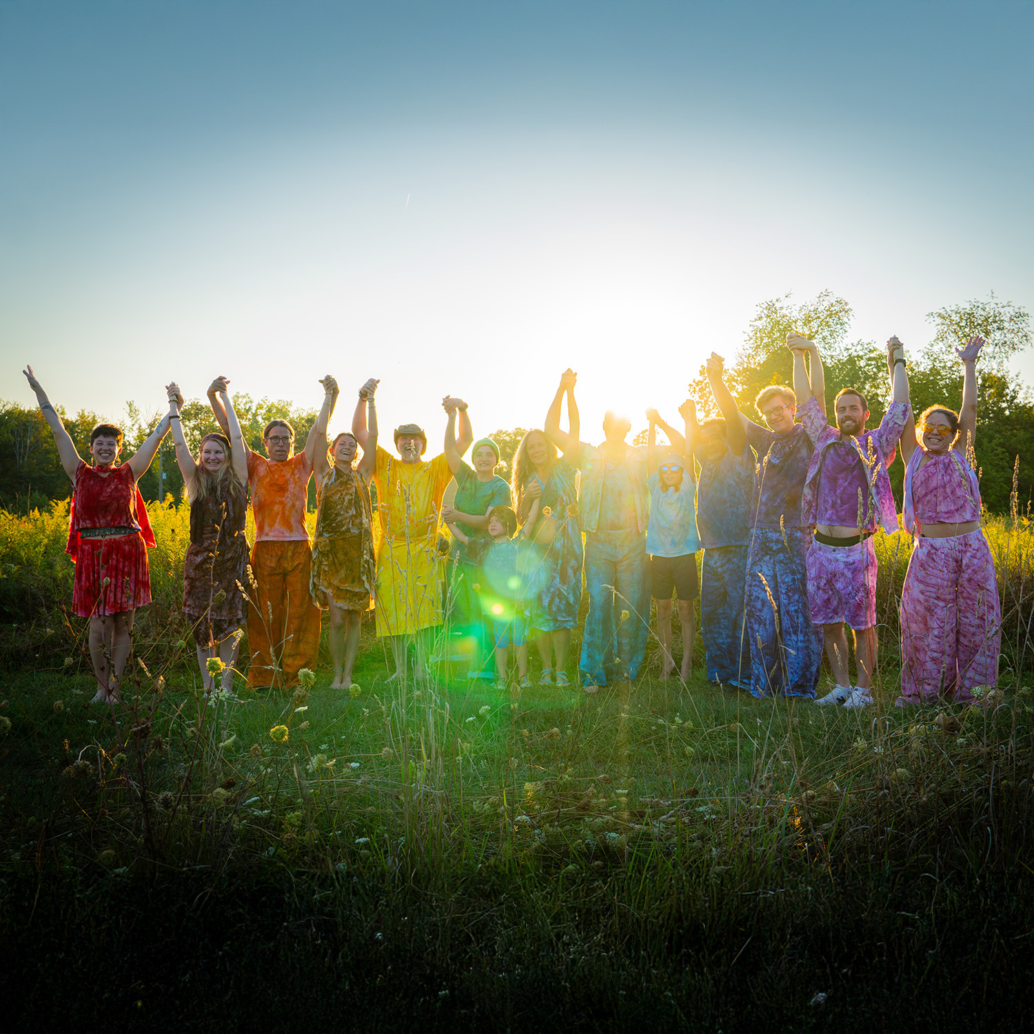 Group of people standing outdoors with arms raised, sunlight shining behind them, in a grassy field with trees.