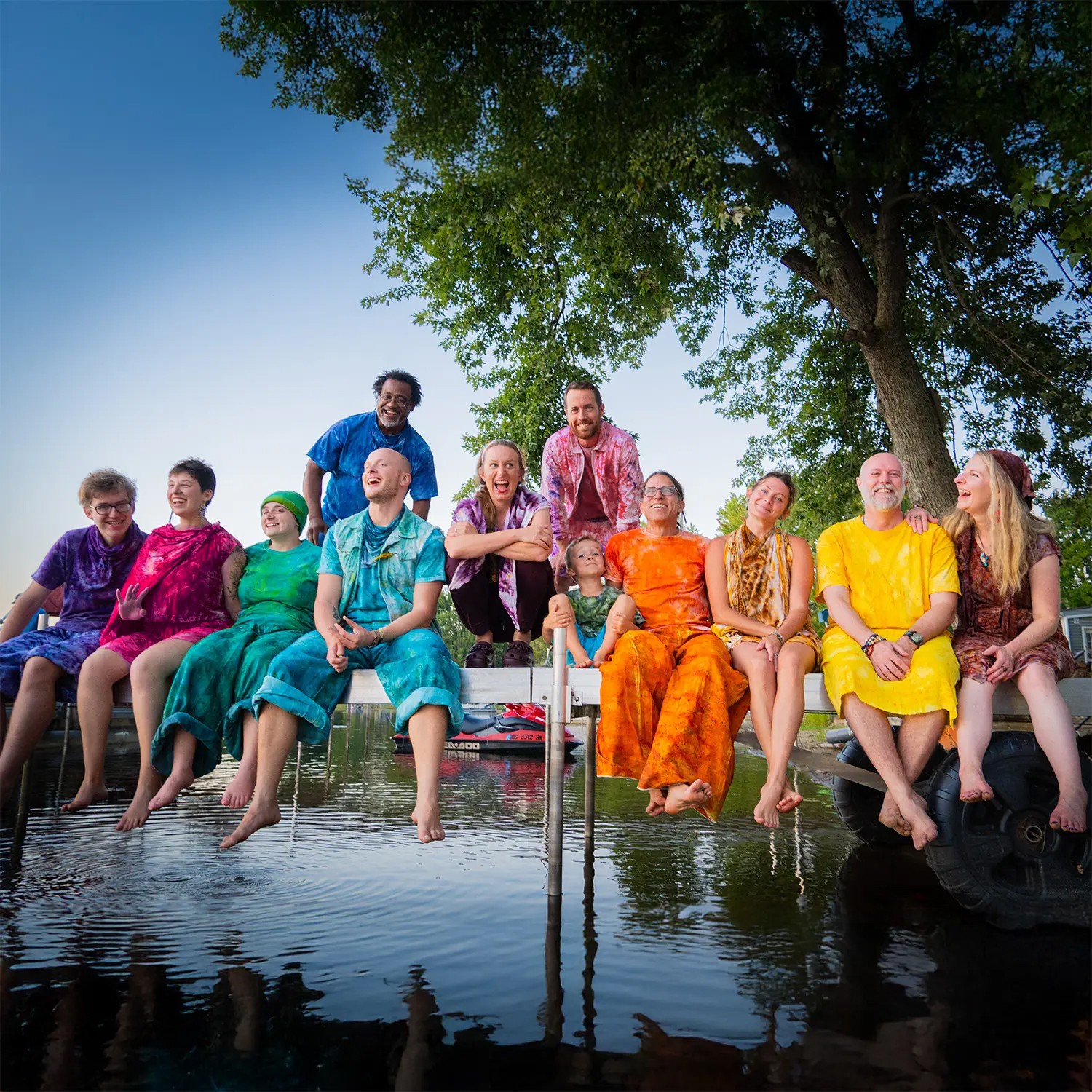 Group of people sitting and standing on a dock with feet in water, under a large tree, sunny day.