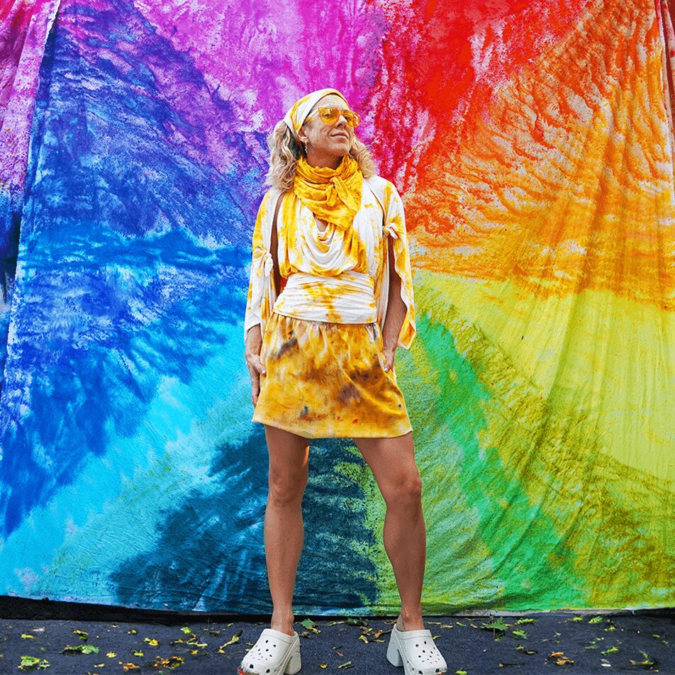 Person standing in front of a colorful tie-dye backdrop, wearing a yellow scarf, skirt, and white shoes.