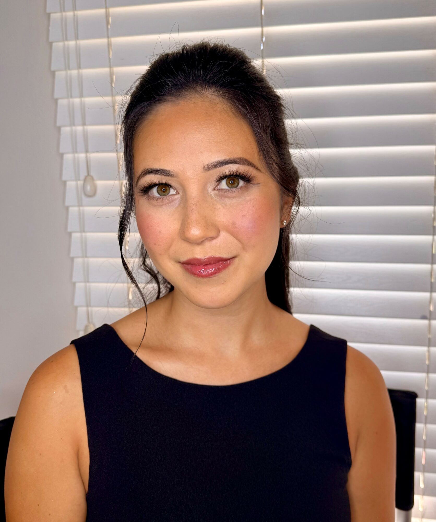 Young woman with dark hair in a ponytail, wearing a sleeveless black top, standing in front of white blinds.