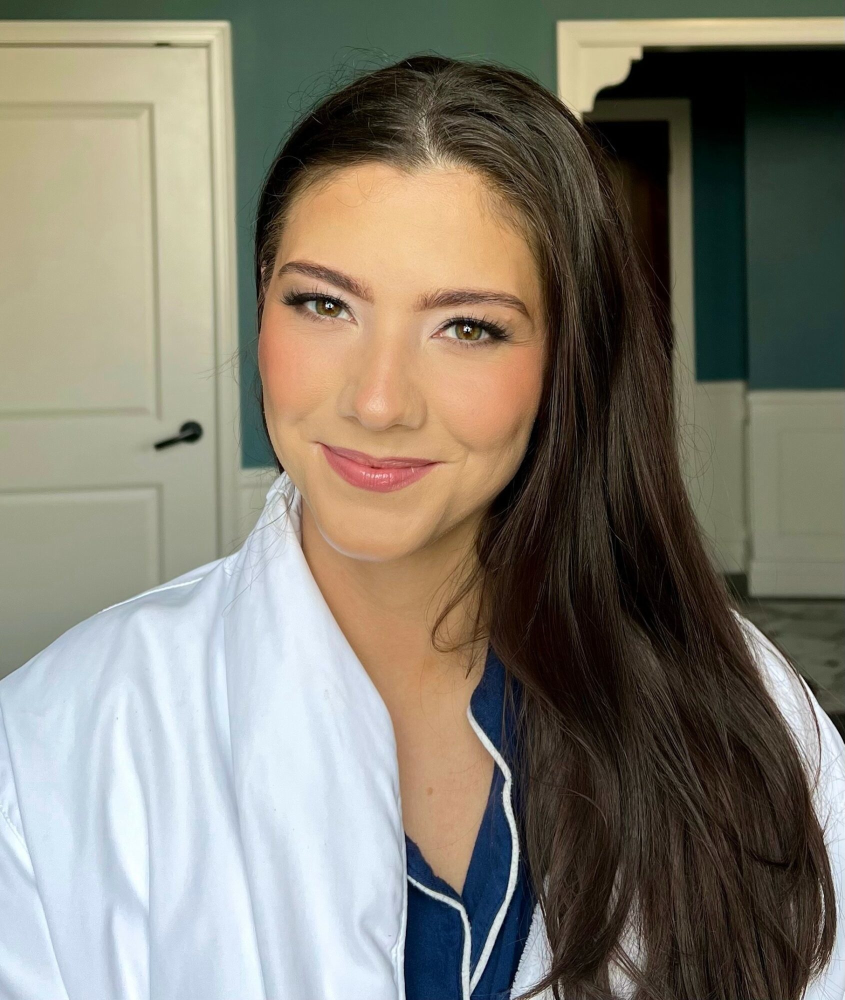 Young woman with long dark hair, smiling, wearing a white shirt, in a room with a door and dark green wall.