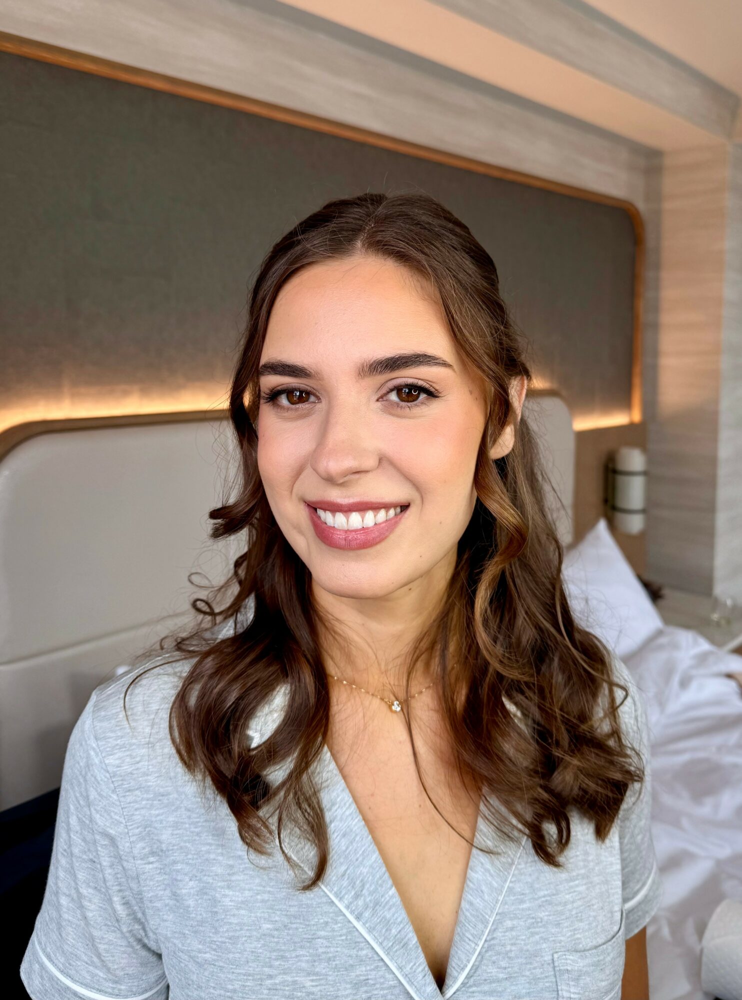 Young woman with wavy brown hair smiling, wearing a light gray top, in a room with a bed and headboard.