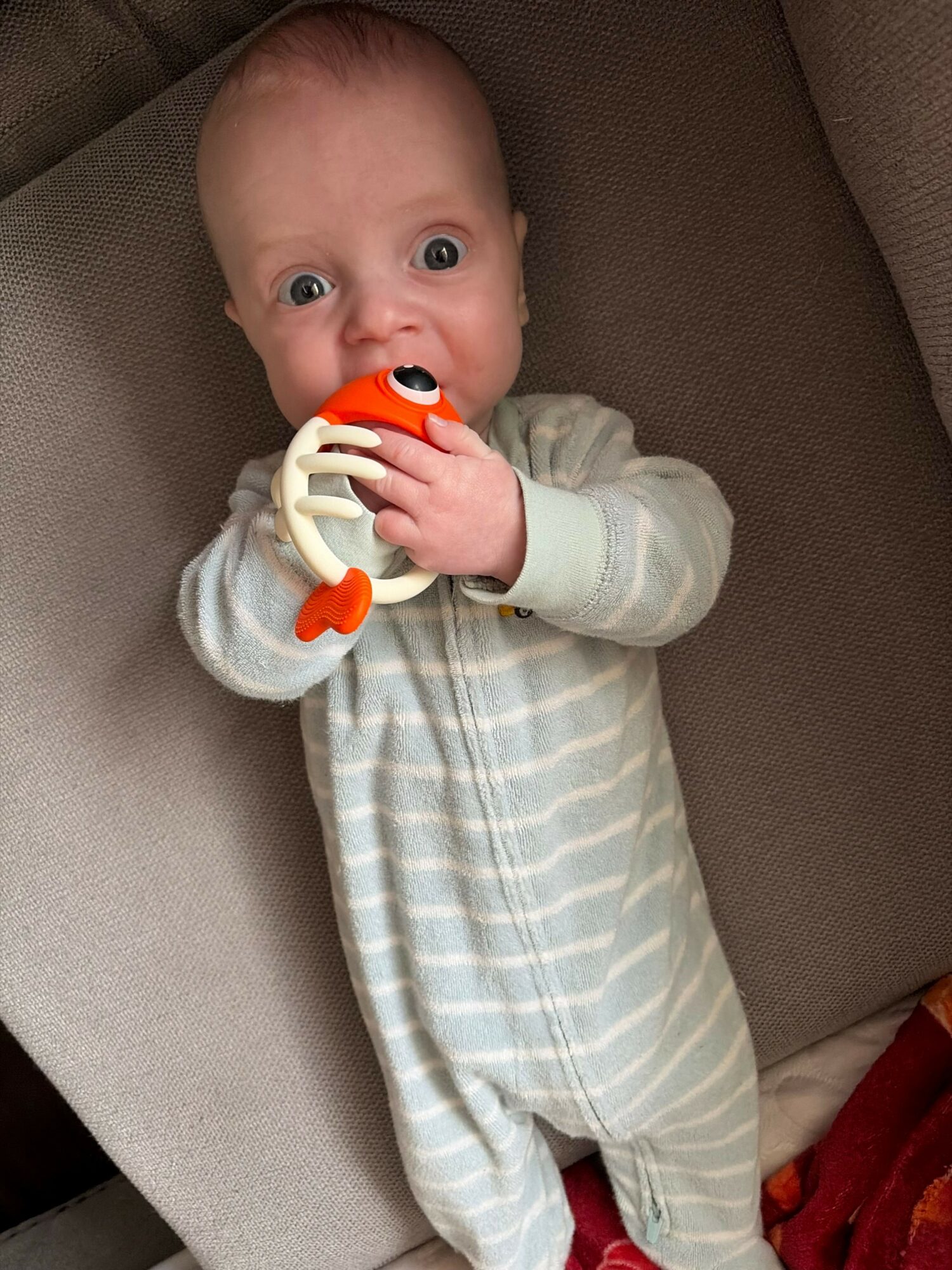 Baby lying on a couch holding a colorful teething toy, wearing a striped onesie, wide-eyed and curious.