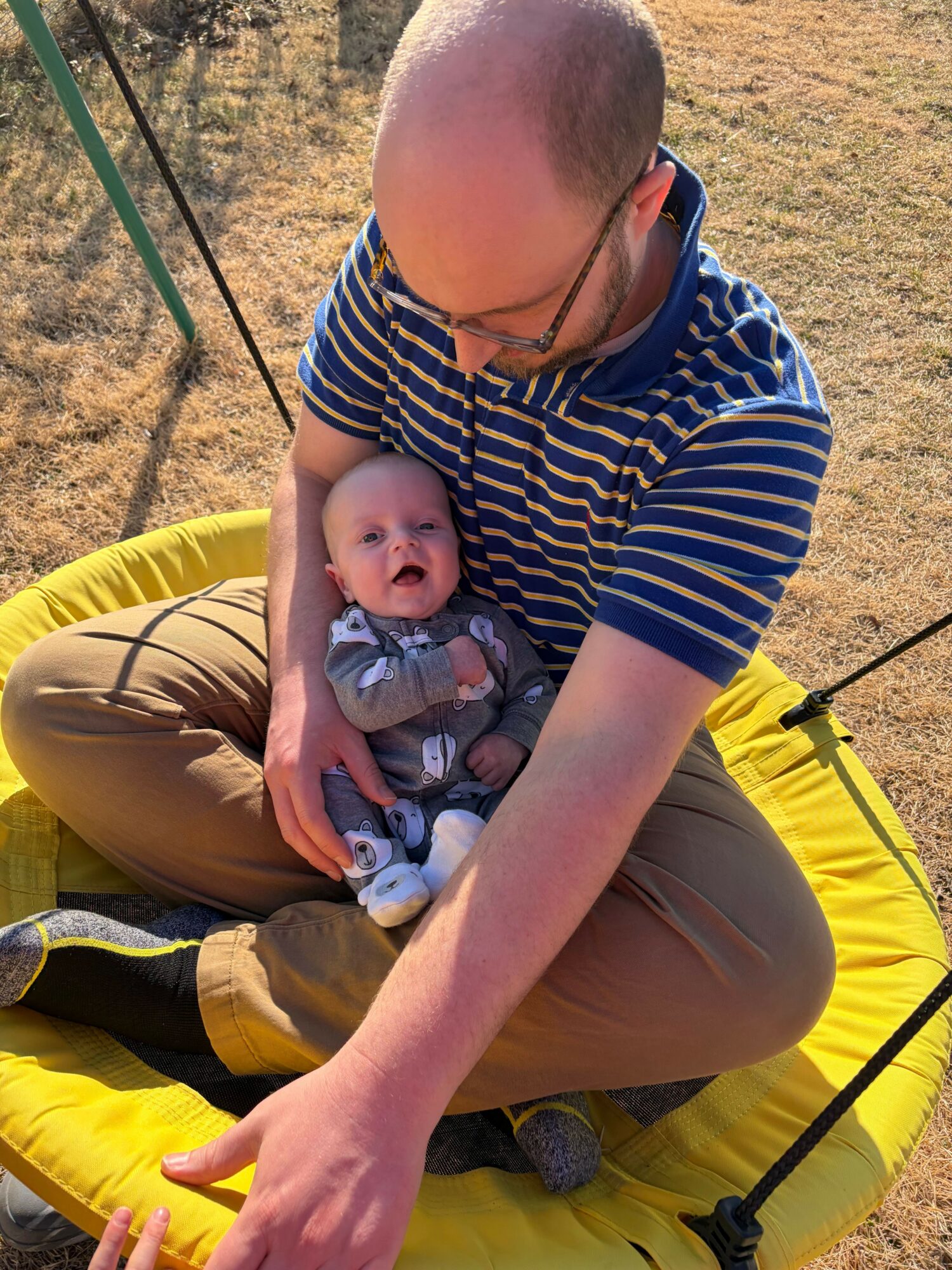 Adult holding a baby on a yellow circular swing outdoors, with grass and trees in background.