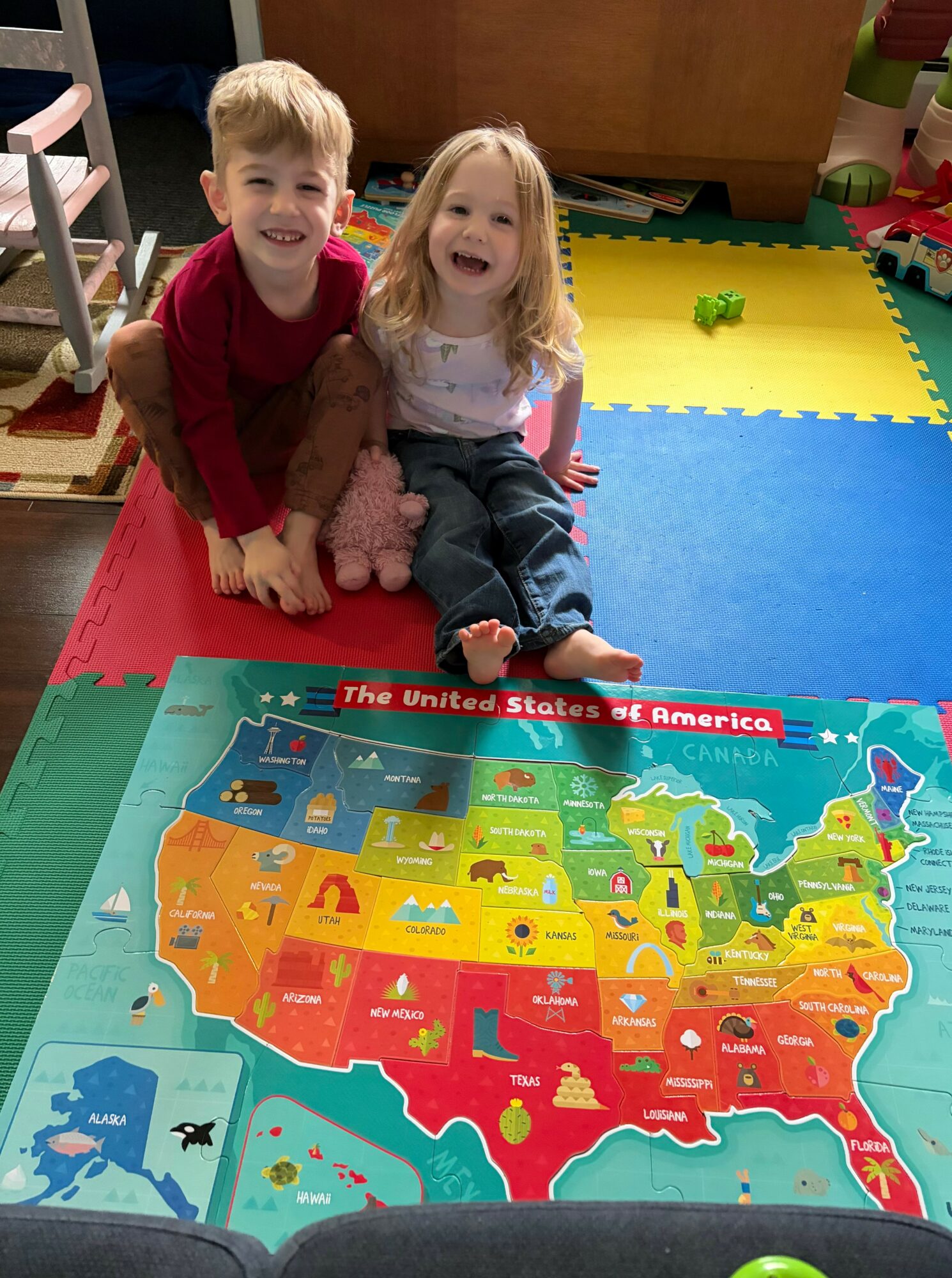 Two children sitting on a colorful foam mat, smiling, with a map of the United States in front of them.