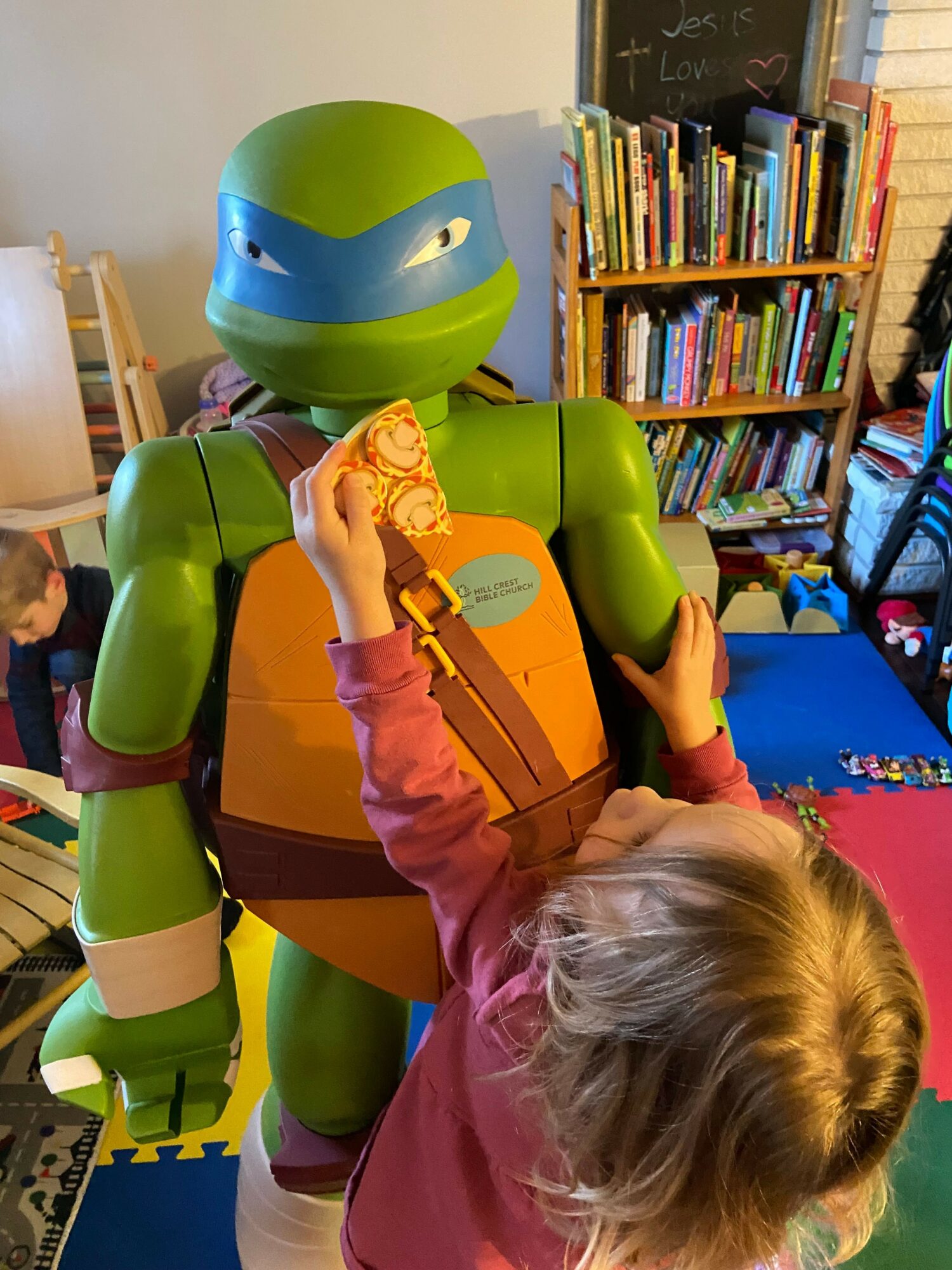 Child with blonde hair interacts with a large green and orange robot toy in a room with bookshelves.
