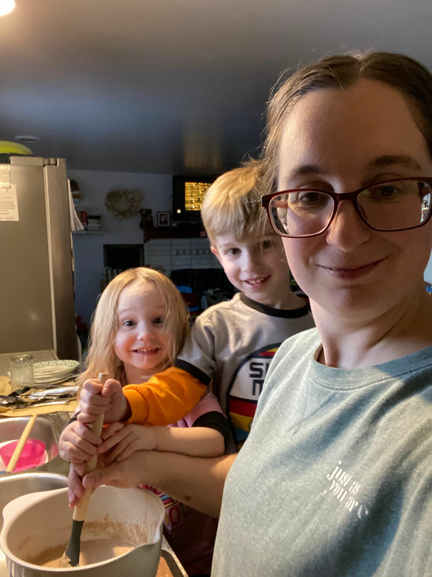 Woman with glasses and two children in a kitchen, one child holding a spoon over a bowl.