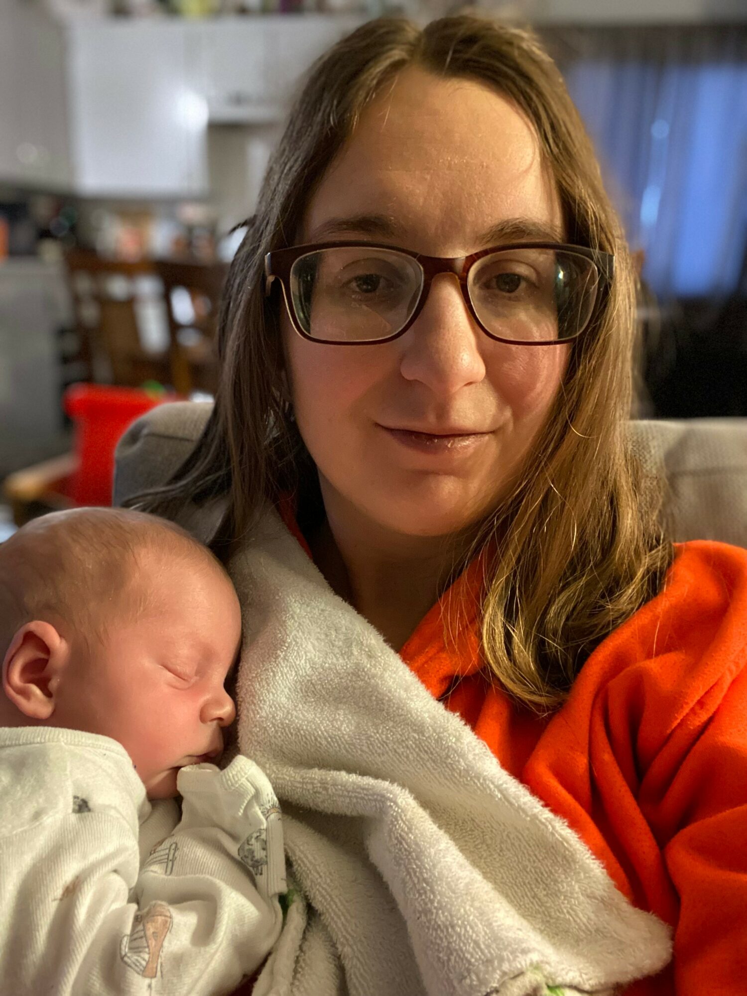 Woman with glasses holding a sleeping baby wrapped in a blanket indoors.