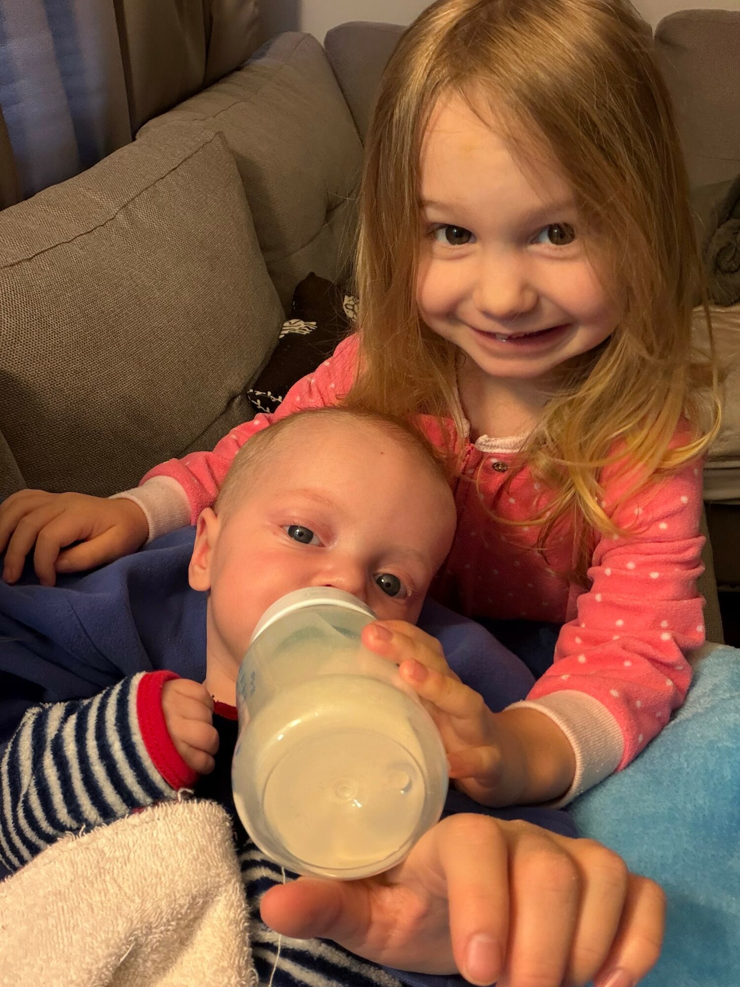 Young girl with long blonde hair smiling, holding a baby drinking from a bottle, sitting on a couch.
