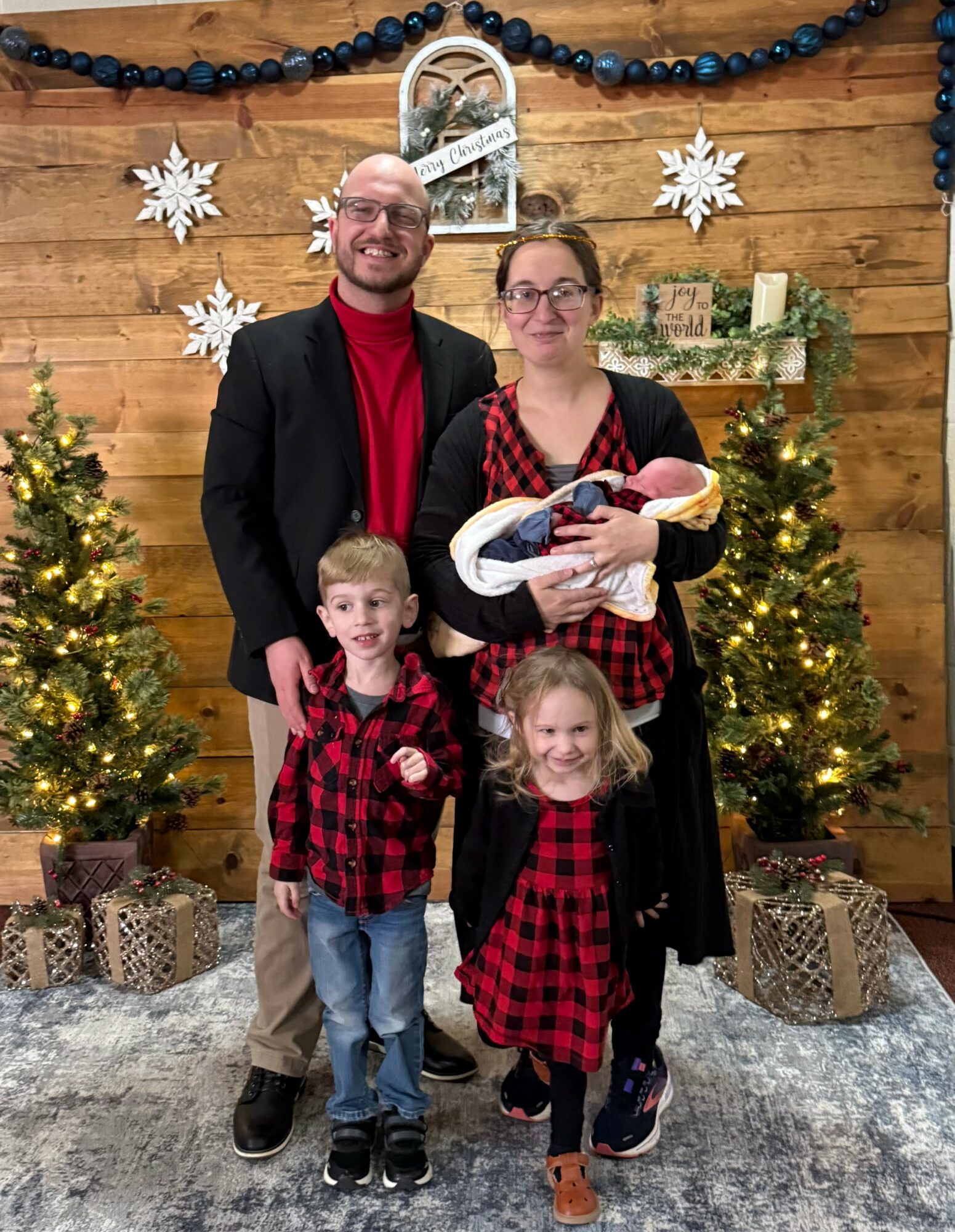 Family of four in Christmas decorations, two children and two adults, smiling, holding a baby, in front of decorated wooden wall.