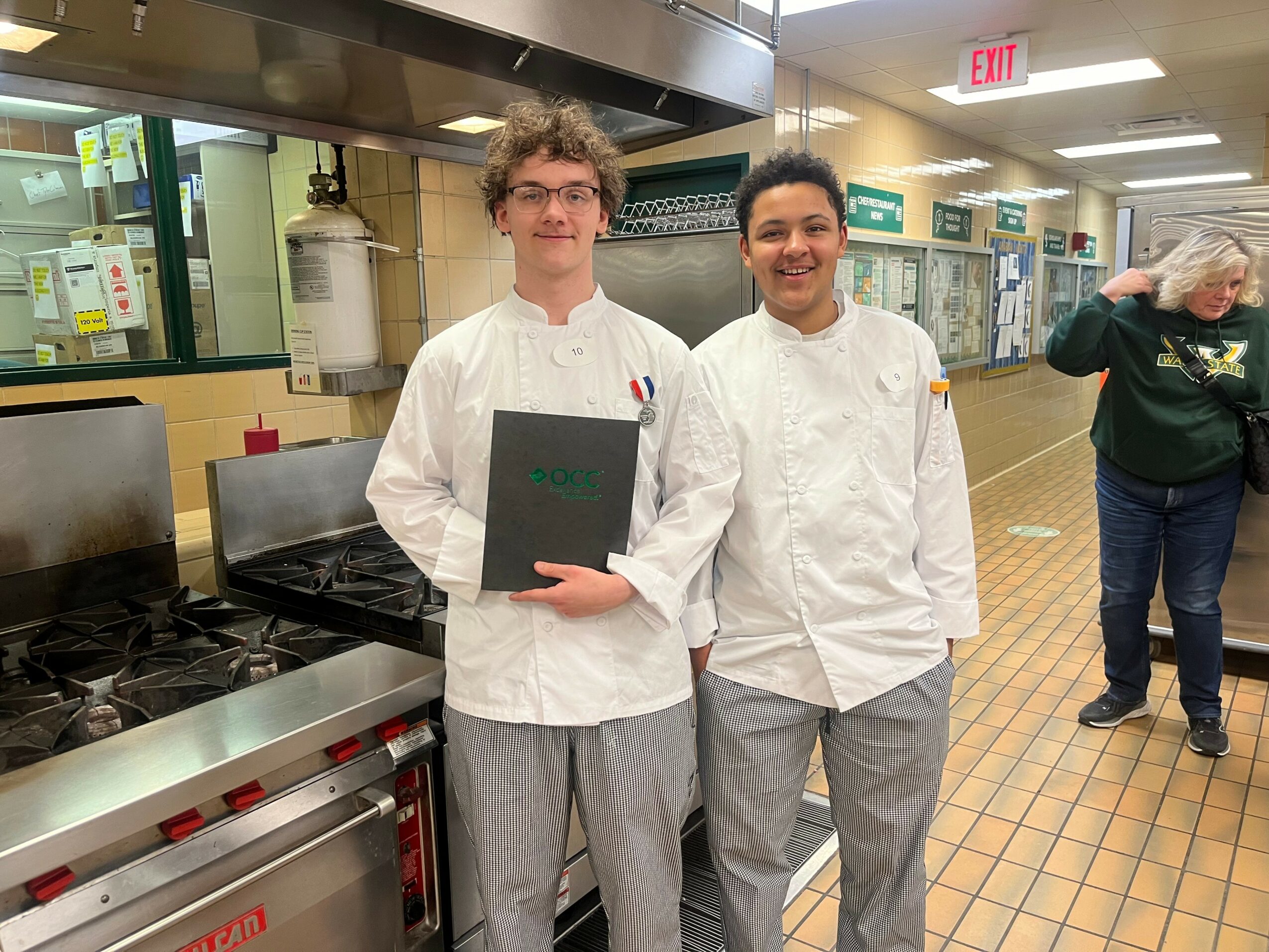 Two young chefs in white uniforms stand in a commercial kitchen, smiling at the camera.