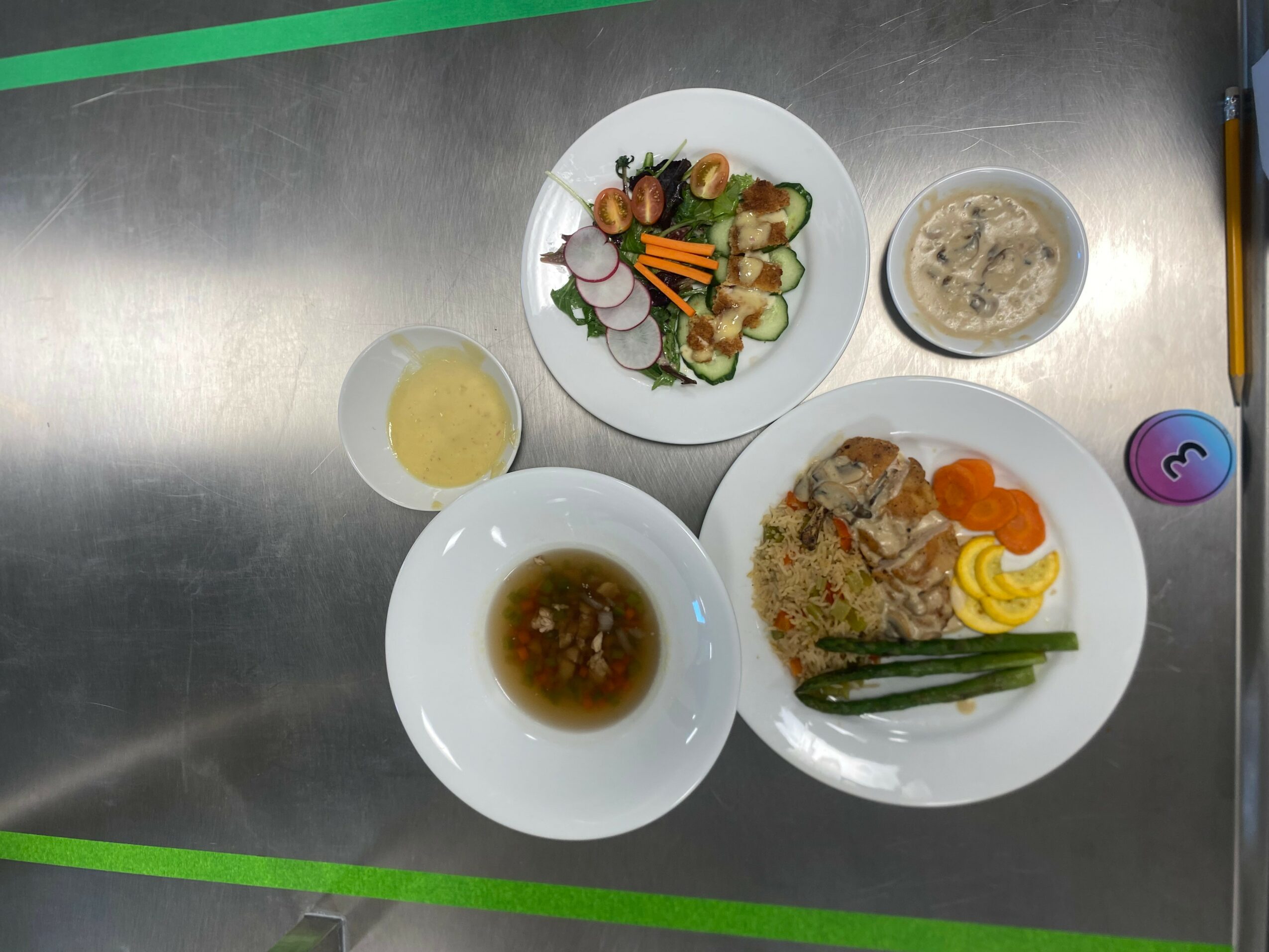 Three plates with food, a bowl of soup, a glass of drink, and a small bowl on a metallic surface.